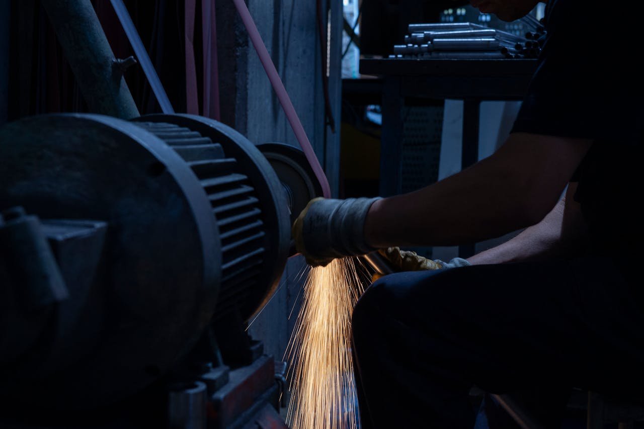 A worker in a factory uses a grinding machine, generating bright sparks in a dark industrial setting.